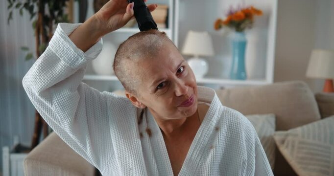 Portrait of a confident and determined woman suffering from cancer, shaving her head with an electric shaver, looking at the camera with a winning smile. Victory over the disease!