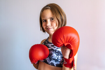 Cheerful young girl in red boxing gloves posing confidently indoors. Promoting children's sport and...