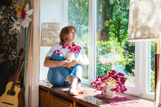 Senior woman sitting on a window seat with Oxalis triangularis plants, enjoying a peaceful moment at home