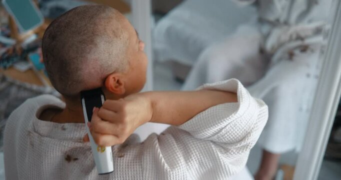 A mature woman with cancer shaves her head with a clipper in front of the mirror at home.