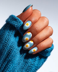 Hand with daisy nail art on matte sky blue nails and cozy knitted sweater
