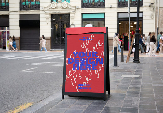 An empty A-frame mockup stands on a busy city sidewalk, ready for advertisements, amidst people walking and shops in the background.