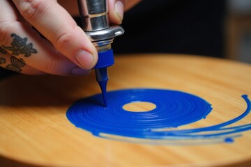 Obraz premium Creating Spiral Art. An artist's hand with a tattoo uses a precision tool to apply blue paint in a spiral on wood.