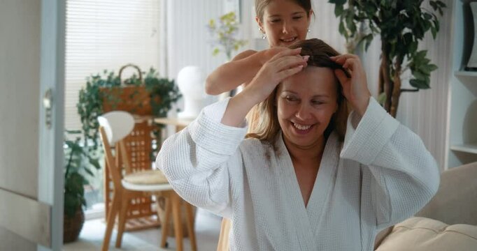 Daughter supports her mom after a haircut, smiling brightly while trying on wigs after chemotherapy in front of a mirror at home. A heartwarming depiction of cancer recovery, self-acceptance, and joy.