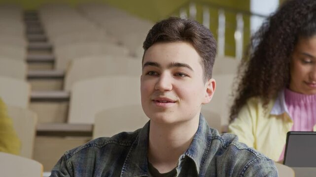 Medium close up shot of young Caucasian guy giving answer and taking notes during lecture with diverse peers in college auditorium