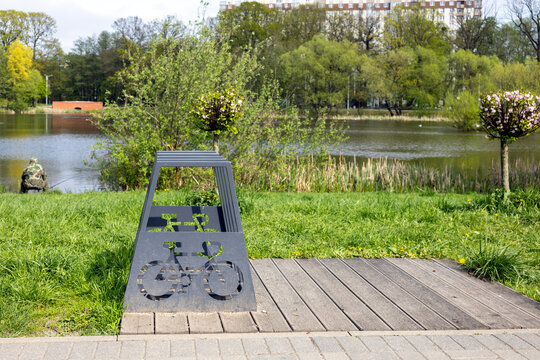 Modern metal bicycle rack on wooden platform near lake in city park with green grass, flowering trees and residential buildings
