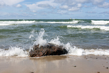 Ocean waves crashing against rock on sandy beach under partly cloudy sky with distant ship on...