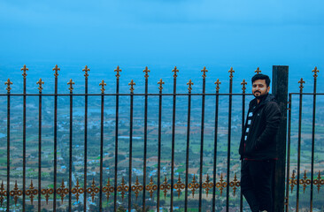 A man in a black jacket leans against a decorative iron fence, gazing at the panoramic valley view...