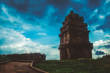 An isolated ancient temple stands against the backdrop of a moody, storm-lit sky at dusk in Badami,...