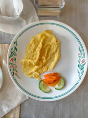 Close up top down view of plain omelette served with tomato and cucumber slices on floral rim white plate. Minimalist breakfast at hotel guesthouse casa particular, Cienfuegos, Cuba. Natural lighting
