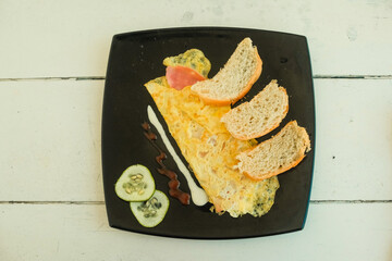 Simple breakfast of omelette, sliced bread, cucumber on black plate on rustic white wooden table. Morning meal in local restaurant, close up top-down view. Travel food photography Varadero, Cuba