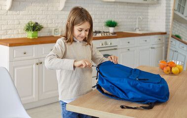 Small pupil boy packing things into blue backpack organizing items before school or learning...