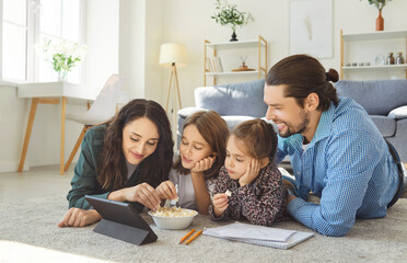 Happy family lying on carpet in living room, watching something on digital tablet and eating popcorn. Smiling parents and children enjoying leisure time, relaxing together during weekend at home.