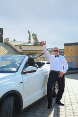 Stylish senior man in bow tie striking fun pose beside white convertible car on rooftop.