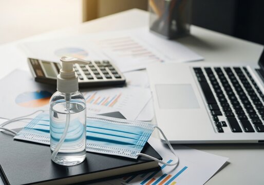 The image depicts a typical office or home office desk setup during the COVID-19 pandemic, featuring work tools alongside hygiene items like hand sanitizer and a face mask