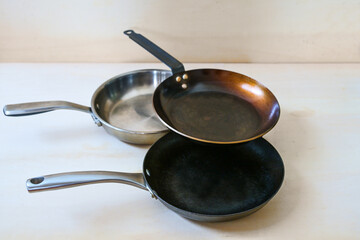 Three frying pans without unhealthy non-stick coating made of carbon steel, stainless steel and cast iron on a light wooden background, copy space, selected focus