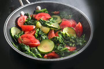Steaming vegetables such as spinach, zucchini and tomatoes in a frying pan on a black stove top, cooking a healthy vegetarian meal at home, selected focus