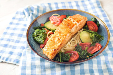 Salmon fillet on spaghetti with spinach, zucchini and tomatoes served in a blue plate on a napkin,...
