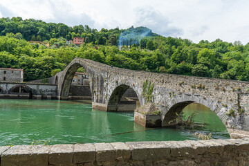Medieval Devil's Bridge crossing green river in Tuscany, Italy, surrounded by lush hills.