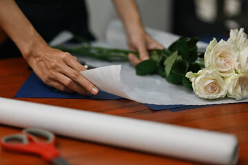 Close-up of florist’s hands wrapping fresh white roses in delicate paper, preparing a bouquet on...