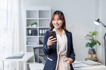Smiling Asian manager holding a smartphone in her hand, standing confidently in her modern office filled with natural light and stylish decor