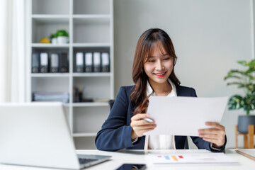 Asian businesswoman sitting at a modern desk, engaging with documents and working on her laptop in...