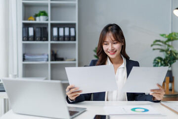 Smiling businesswoman reading and comparing two documents while sitting at her desk in a modern...