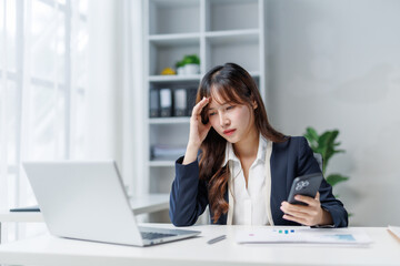 Young Asian businesswoman is having headache while using smartphone and working with laptop at office