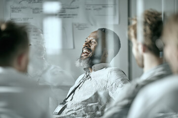 A black man in his late thirties with short hair and beard, laughing while leading an office meeting around whiteboard with blurred figures of male team members looking at the board. 
