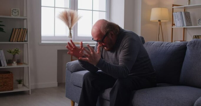 Frustrated elderly man sitting on couch at home, expressing anger, grief or distress with raised hands. Desperate senior male struggling with emotional pain, despair or blaming himself for mistakes.