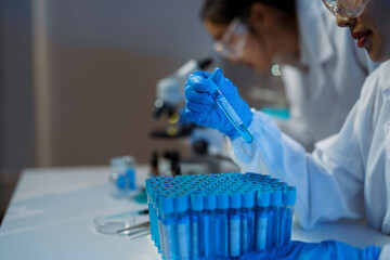 Scientist wearing gloves analyzing blue liquid in test tube with microscope in background in...