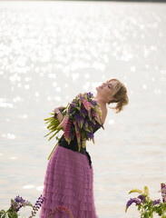 A girl in a beautiful dress walks on the shore of a lake with a bouquet of lupines