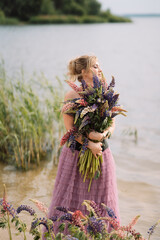 A girl in a beautiful dress walks on the shore of a lake with a bouquet of lupines