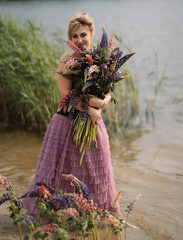 A girl in a beautiful dress walks on the shore of a lake with a bouquet of lupines