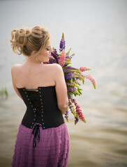A girl in a beautiful dress walks on the shore of a lake with a bouquet of lupines