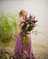 A girl in a beautiful dress walks on the shore of a lake with a bouquet of lupines