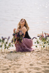 A girl in a beautiful dress walks on the shore of a lake with a bouquet of lupines