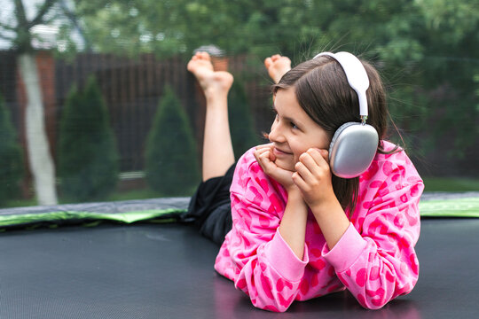 A cheerful child girl 8-9 years in a pink leopard print sweatshirt with headphones lying on a trampoline and smiling, concept of childhood and relaxation, copyspace