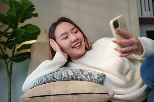 Happy Asian woman relaxing on a cozy sofa, using her smartphone while enjoying leisure time in a modern living room