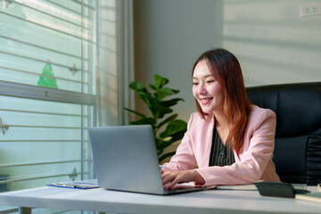 Smiling Asian businesswoman working on a laptop in a festive office, surrounded by holiday decorations and a Christmas tree