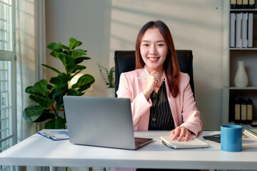Young manager working at her desk, smiling brightly while engaging with the camera, surrounded by a laptop and important documents
