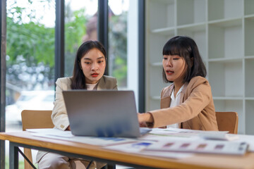 Fototapeta premium Two Asian businesswomen collaborating and discussing strategies while working on a laptop in a modern office environment