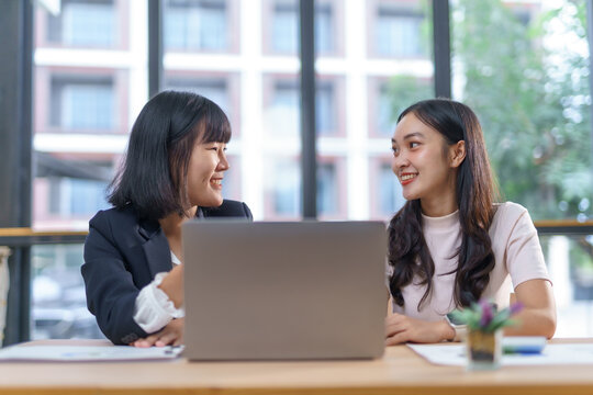 Two Asian businesswomen discussing work while using a laptop in a modern office setting, showcasing collaboration and professional communication
