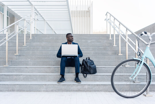 African american Businessman or student working on laptop while sitting on steps with bicycle nearby