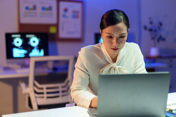 Asian businesswoman working late at night using laptop in office, concentrating on her tasks with determination