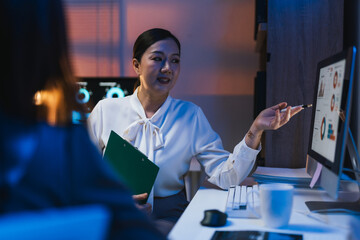 Asian businesswoman holding a clipboard and pointing at computer screen showing charts while explaining project to her colleague during a late night meeting