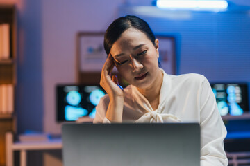 Exhausted medical worker having a bad headache while working on her laptop at night, with monitor showing brain scans in the background