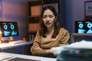 Tired and worried businesswoman looking down with arms crossed, working with financial data on computer screen late at night in office