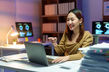 Young professional having a video conference on laptop at her desk in the office late at night, pointing her finger at the screen and smiling