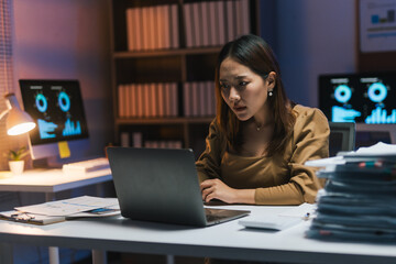 Focused businesswoman working late using laptop in office at night, graphs showing on computer screens
